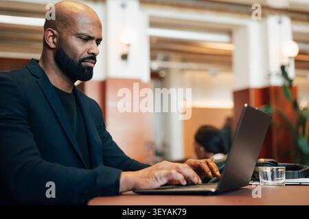 Ein fokussierter Geschäftsmann, der auf seinem Laptop Post schreibt, während er in einem modernen Coworking-Bereich sitzt und arbeitet Stockfoto