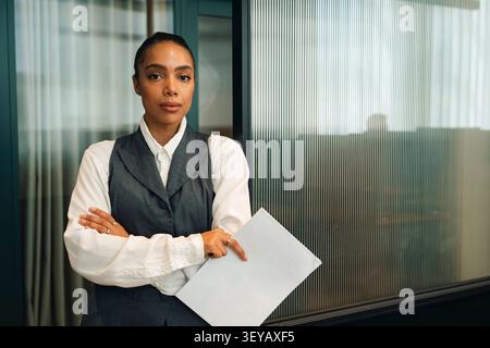 Porträt einer selbstbewussten Geschäftsfrau, die ein Dokument hält, während sie im Büro mit überkreuzten Armen posiert Stockfoto
