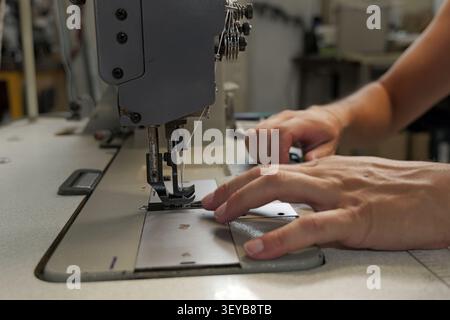 Hände eines Bekleidungsarbeiters, der eine industrielle Nähmaschine bedient, führen den Stoff sorgfältig durch die Maschine und demonstrieren das Handwerk der Schneiderei Stockfoto