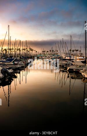 Zahlreiche Segelboote liegen während eines farbenfrohen Sonnenuntergangs in einem ruhigen Hafen vor, deren Masten auffällige Reflexionen auf der stillen Wasseroberfläche erzeugen Stockfoto
