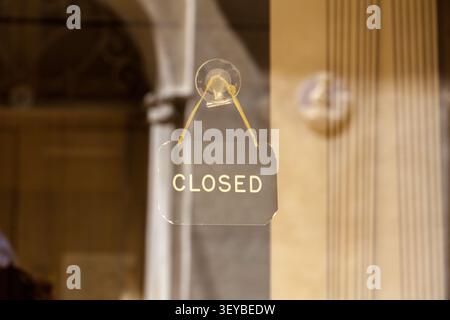 Ein dunkles Glasschild „Geschlossen“ hängt an einem Saugnapf in einem Schaufenster, mit verschwommenen Steinsäulen im Hintergrund, Gozo Stockfoto