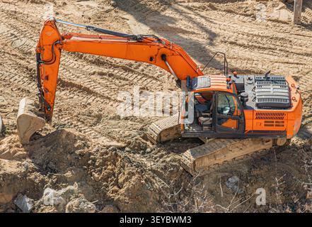 Orangefarbener Bagger im Einsatz auf schlammiger Baustelle, Arm ausgefahren bei abgesenkter Schaufel, Raupenketten sichtbar, Kabine geschlossen, Modellnummer DX200A angezeigt, Stockfoto