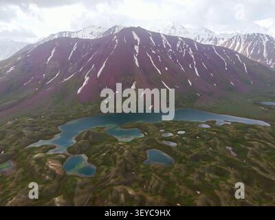 Panoramablick auf einen langen türkisfarbenen Bergsee unter dem einzigartigen rötlichen Schichtschlamm und den Schneegipfeln Stockfoto