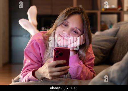 Eine junge Frau mit langen Haaren beschäftigt sich fröhlich mit ihrem rosa Smartphone. Sie trägt ein einfaches rosa T-Shirt und liegt auf der Couch im Wohnzimmer Stockfoto