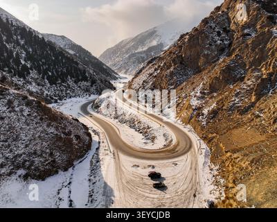 Aus der Vogelperspektive auf eine kurvenreiche asphaltierte Straße mit einer scharfen Haarnadelkurve in einer zerklüfteten, schneebedeckten Bergschlucht mit steilen, trockenen Hängen Stockfoto
