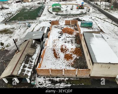 Von oben aus der Vogelperspektive auf ein Baufundament, einen Kunstrasen-Fußballplatz und einen farbenfrohen Kinderspielplatz in einem schneebedeckten Vorort Stockfoto