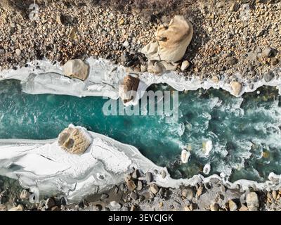 Blick von oben auf den pulsierenden türkisfarbenen Gletscherfluss, der zwischen dicken Schichten von weißem Eis und steilen, felsigen, bunten Ufern fließt Stockfoto