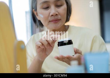 Schöne reife asiatische alte Frau, die Hautpflegeprodukte in einer Schönheitsroutine aufträgt. Selektiver Fokus. Gesundes Pflegekonzept genießen. Stockfoto