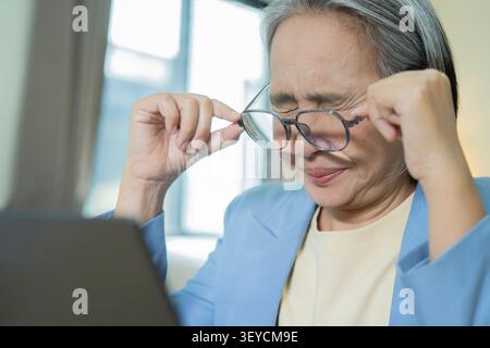 Reife Frau fühlt Kopfschmerzen und Stress, während sie im Home Office an einem Laptop arbeitet. Müde Angestellte mit geschlossenen Augen schrubbt den Tempel, zeigt Müdigkeit und Geisteskrankheit Stockfoto