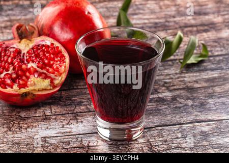 Leuchtender roter Granatapfelsaft in klarem Glas neben halbem Obst auf verwittertem Holz. Satter Farbkontrast, natürliches Licht, geringe Schärfentiefe. Evo Stockfoto