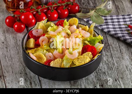 Frischer Caesar-Salat mit Garnelen, Croutons, Kirschtomaten, Salat in schwarzer Schüssel. Rustikaler Holztischhintergrund mit karierter Serviette. Warmes Licht Stockfoto