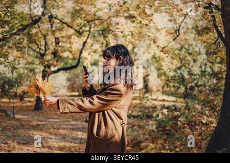 Die Person nimmt ein Smartphone-Foto von gelben Herbstblättern auf, während sie im Park lächelt. Social Media Influencer Lifestyle, Herbst Bloginhalte, Lifestyle Stockfoto