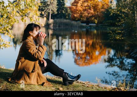 Die Frau sitzt im Herbstpark auf dem Grasufer am Teich und trinkt aus dem Becher. Spirituelle Verbindung mit der Natur, Rückansicht Alleinreisender, achtsame Erholung Stockfoto
