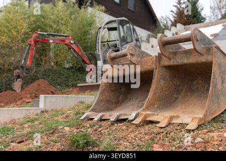Nahaufnahme von rostigen Baggerlöffeln, die auf einer Baustelle auf dem Boden liegen. Ein Minibagger und ein Privathaus sind im Hintergrund zu sehen Stockfoto