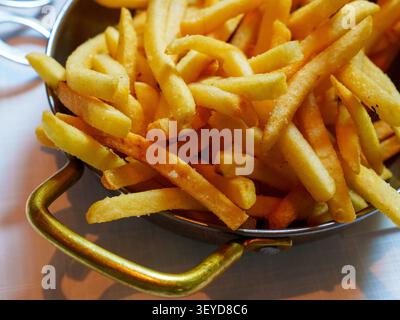 Aus nächster Nähe sehen Sie salzige, goldgebratene Kartoffelstäbchen, die in einer Metallpfanne mit verzierten Messinggriffen serviert werden, und laden zu zwanglosen Speisen ein. Stockfoto