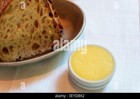 Zwei Scheiben handwerkliches Sauerteigbrot, serviert in einer Vintage-Metallschale, zusammen mit einem kleinen Ramekin aus glatter gelber Butter auf einer weißen Tischdecke. Stockfoto