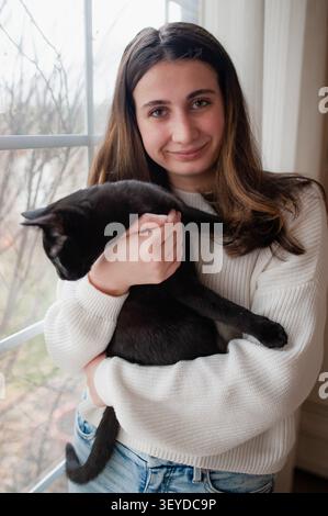 Lächelnde junge Frau, die eine schwarze Hauskatze im Pullover am Fenster hält Stockfoto