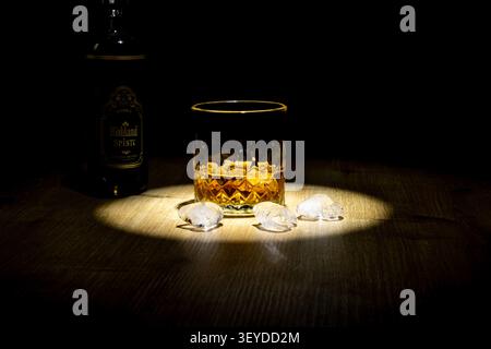 Dramatisches Stillleben eines Whiskyglases mit goldenem Bernsteinbrand und Eiswürfeln auf einer dunklen Holzoberfläche. Eine Whiskyflasche mit Elega Stockfoto