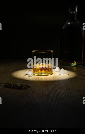 Stimmungsvolles Stillleben mit einem Whiskyglas im Kristallschliff mit goldenem Bernsteinbrand und Eiswürfeln auf einer dunklen Holzoberfläche. Eine Glasflasche steht rein Stockfoto