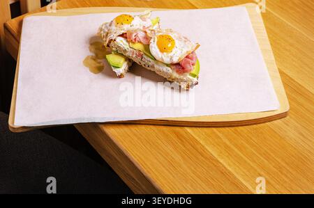 Zwei Stücke herzhafter Avocado Toast mit Spiegeleiern und Schinken auf einem Holztisch Stockfoto