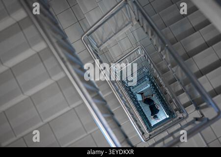 Ein dramatischer Blick von oben auf eine Person, die allein am Fuß einer modernen Treppe liegt. Die geometrische Architektur unterstreicht Themen der Isolation Stockfoto