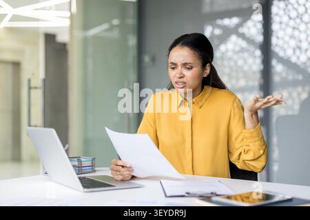 Frustrierte Geschäftsfrau, die sich ein Dokument ansieht, während sie mit den Schultern zuckt, sich durch ein Problem oder einen Fehler verwirrt und gestresst fühlt und in ihrem modernen Büro arbeitet Stockfoto