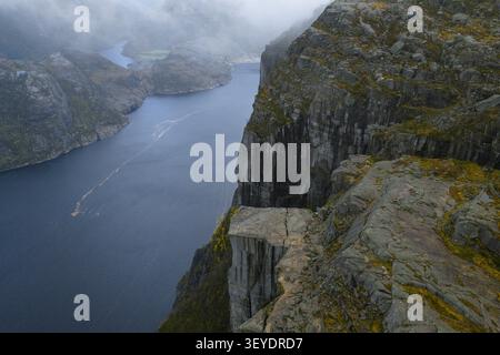 Aus der Vogelperspektive der berühmten Preikestolen Klippe, die über dem tiefblauen Wasser des Lysefjords mit nebeligen Wolken und zerklüftetem felsigem Gelände in Songesand thront. Stockfoto