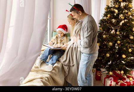 Glückliche Familie, die Weihnachten genießt, Kinder mit Buch auf der Fensterbank lesen Stockfoto