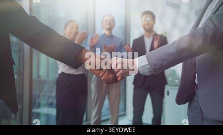 Nahaufnahme kaukasische und afrikanische Geschäftsleute schütteln die Hände in der Nähe von unbeschwerten drei multiethnischen Partnern, die fröhlich im sonnigen Firmenbüro applaudieren Stockfoto