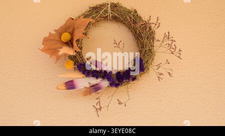 Fallkranz aus Stroh hängt an einer strukturierten Wand. Handgefertigter Herbstkranz mit lila Blumen, Federn und getrockneten Blättern. Handgefertigte saisonale Dekoration Stockfoto