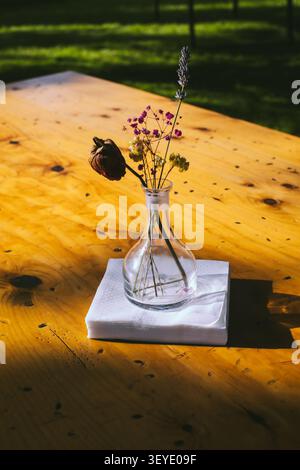 Anordnung von getrockneten Blumen in einer klaren Glasvase auf einem gefalteten weißen Serviette auf einem Holztisch mit natürlichem Licht Stockfoto