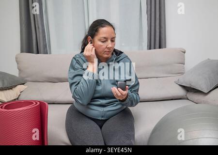 Eine konzentrierte Frau, die einen kabellosen Ohrstöpsel einsetzt, bevor sie mit ihrem Fitness-Training zu Hause beginnt, und auf einem Sofa sitzt, mit Trainingsgeräten in der Nähe. Stockfoto