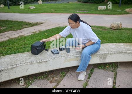 Frau, die auf einer Steinbank sitzt und Drohnenausrüstung vorbereitet, Batterien und Controller auf der Bank in einem grünen Park platziert. Stockfoto