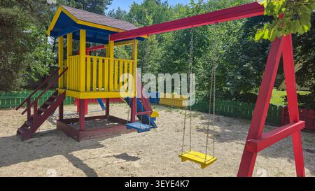 Heller, farbenfroher Holzspielplatz mit Spielhaus, Schaukel, Rutsche und Sandkasten, umgeben von grünen Bäumen in einem sonnigen Park. Freizeitangebot für Kinder im Freien Stockfoto