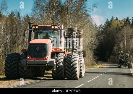 Großer roter knickgelenkter Traktor mit Kultivator auf Asphaltstraße. Roter Kirovets-Traktor mit eingeklapptem Grubber, der auf einer ländlichen Asphaltstraße fährt Stockfoto