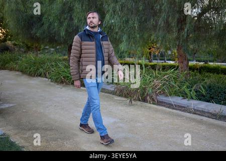 Ein Mann in einer warmen Jacke, der an einem kalten Herbsttag einen Park entlang geht, umgeben von Grün. Stockfoto