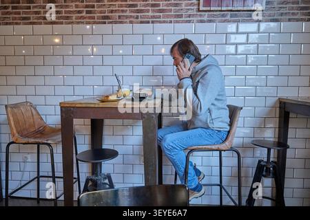 Mann, der im Café arbeitet, während er mit dem Smartphone spricht und mit dem Laptop arbeitet. Stockfoto