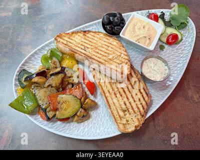 Italienische Antipasti-Platte mit gegrilltem Brot, geröstetem Gemüse, Oliven und Dips, serviert auf einer Keramikplatte. Stockfoto