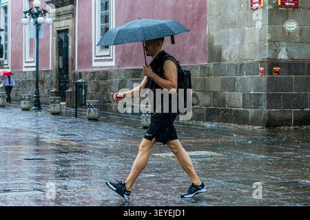 Ein Mann in Sportkleidung mit einem Rucksack geht durch einen starken Regenschauer und hält einen Regenschirm auf den Straßen von Puebla. Stockfoto