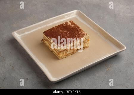 Klassisches Tiramisu-Dessertquadrat auf Keramikplatte mit Kakaostaubung auf rustikaler Steinoberfläche Stockfoto