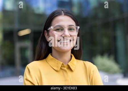 Junge, selbstbewusste Frau, die eine klare Brille und ein gelbes Hemd trägt, lächelt breit in die Kamera, während sie vor einem modernen Bürogebäude steht Stockfoto
