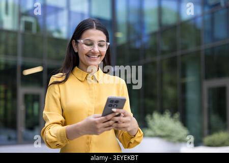 Junge lächelnde Frau mit Brille, die vor einem modernen Bürogebäude steht, ein Smartphone in der Hand hält und sich aktiv mit der mobilen Technologie beschäftigt Stockfoto