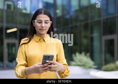 Junge Geschäftsfrau mit Brille und gelbem Hemd. Im Freien stehen und aktiv mit ihrem Smartphone interagieren. Repräsentiert moderne Kommunikation. Business-Konnektivität Stockfoto