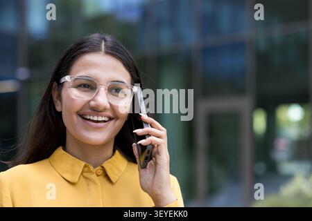 Junge indianerin mit Brille und gelbem Hemd, die sich vor einem Bürogebäude glücklich auf einem Mobiltelefon unterhalten, was für geschäftliche Kommunikation und beruflichen Erfolg steht Stockfoto
