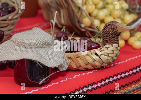 Hausgemachte Kirschmarmelade, serviert in einem rustikalen Glasglas mit frischen süßen Kirschen, gewebtem Korb und traditionellem rotem Textil, die saisonale Ernte feiert Stockfoto