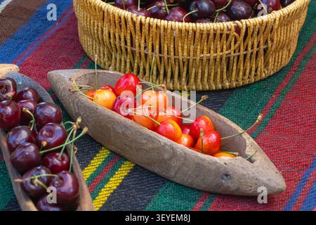 Frische süße Kirschen in einer traditionellen geschnitzten Holzschale mit rustikalem Korb auf farbenfroher bulgarischer Volkstextilien für eine authentische Ernte Stockfoto
