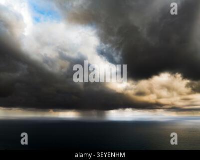 Eine riesige Weite dunkler, stimmungsvoller Wolken erhebt sich über dem tiefblauen Meer, mit einem sichtbaren Regenschauer, der am Horizont herabfällt Stockfoto
