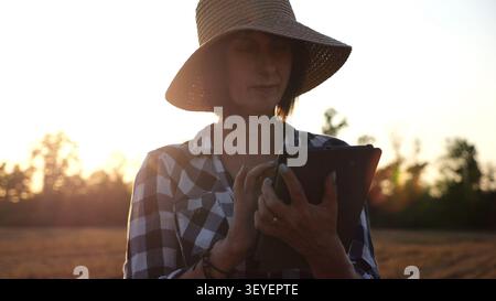 Agrostomistin im Strohhut, die die Ernte auf dem Gerstenfeld bei Sonnenuntergang überwacht. Erwachsener Landwirt mit digitaler Tablette auf Weizenwiese in der Abenddämmerung. Schön Stockfoto
