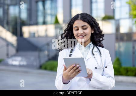 Ärztin in Laborkittel und Stethoskop lächelt, während sie ein digitales Tablet benutzt, steht außerhalb eines modernen Gebäudes und steht für Innovation und Konnektivität im Gesundheitswesen Stockfoto