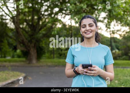 Junge Frau lächelt und hält ein Smartphone mit Ohrhörern, macht eine Pause von ihrem Workout in einem grünen Park und repräsentiert Fitness, Wellness und einen aktiven Lebensstil Stockfoto